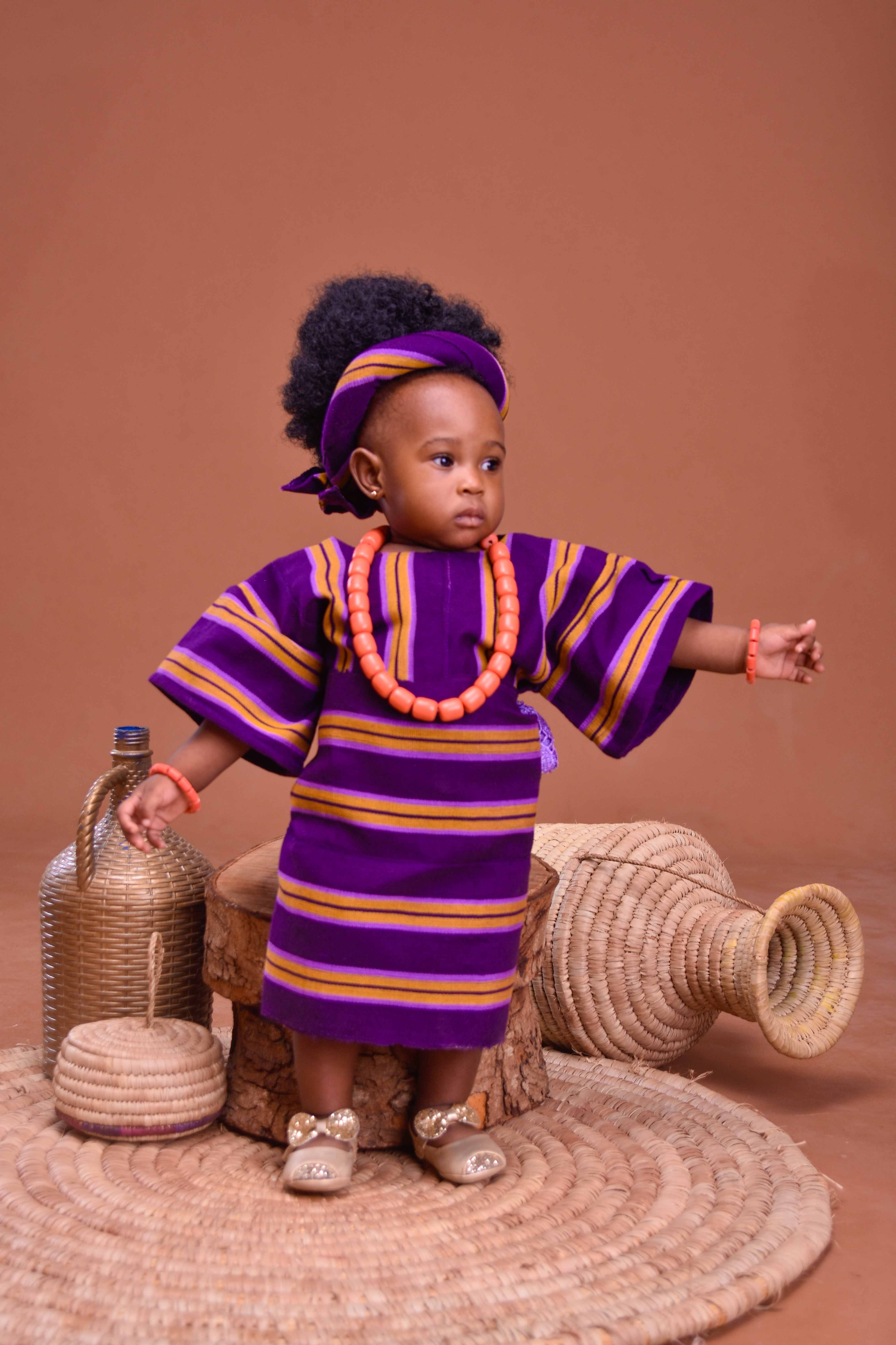 A baby in african clothing standing next to a basket  Free Stock Photo