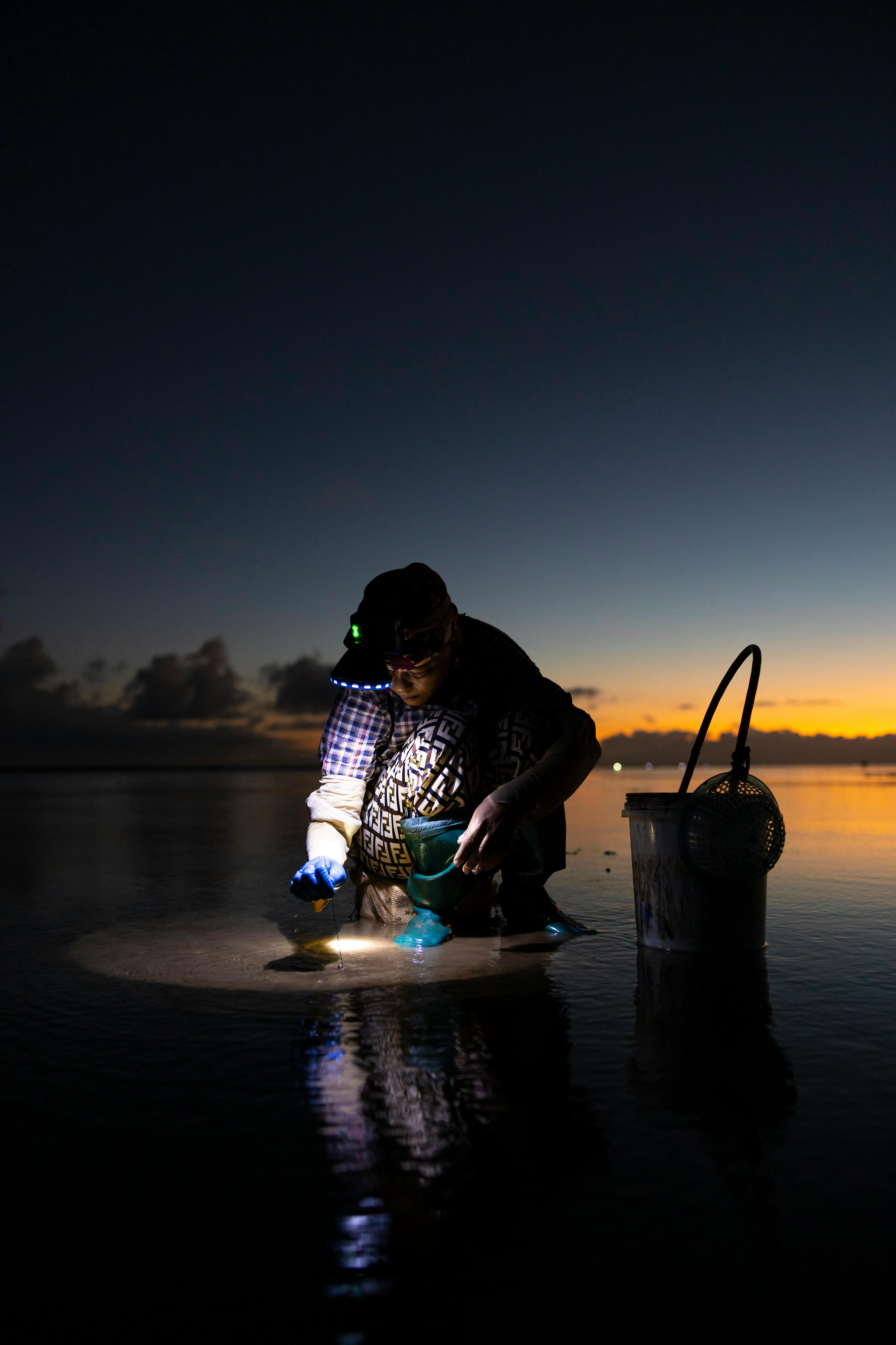 A person collects shells by flashlight on a calm beach at twilight, reflecting on serene waters.