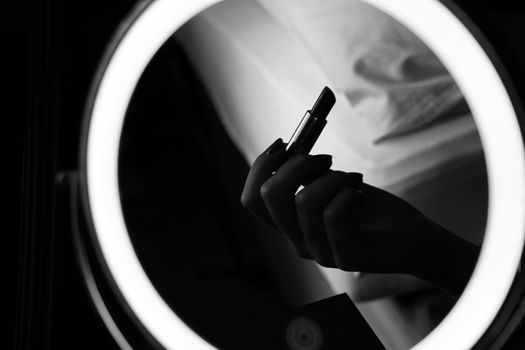 Artistic black and white photo of a woman holding lipstick in front of vanity mirror.
