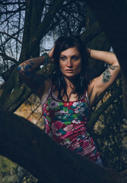 Free Portrait of a tattooed woman posing confidently in a floral dress amidst tree branches. Stock Photo