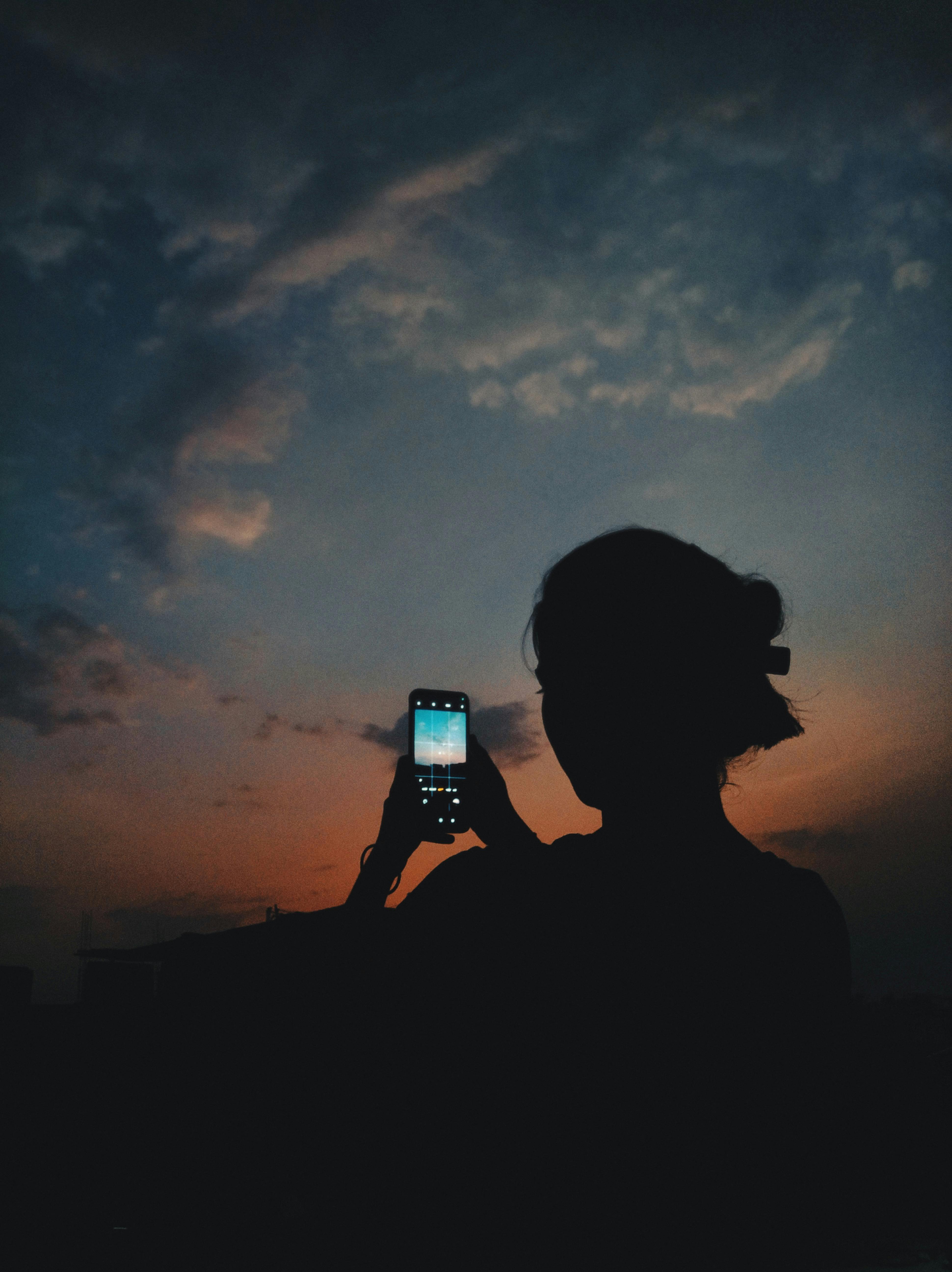 Silhouette of a woman holding a phone, capturing a vibrant sunset sky.