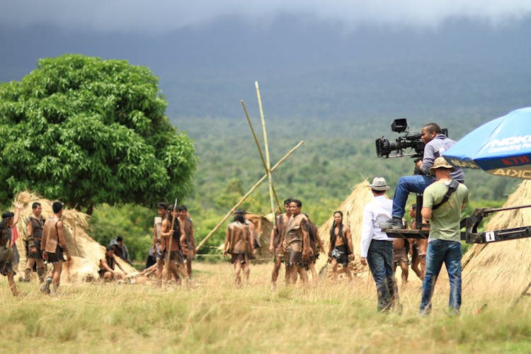 People At The Green Grass Field With The Distance Holding Filming Camera During Day Time