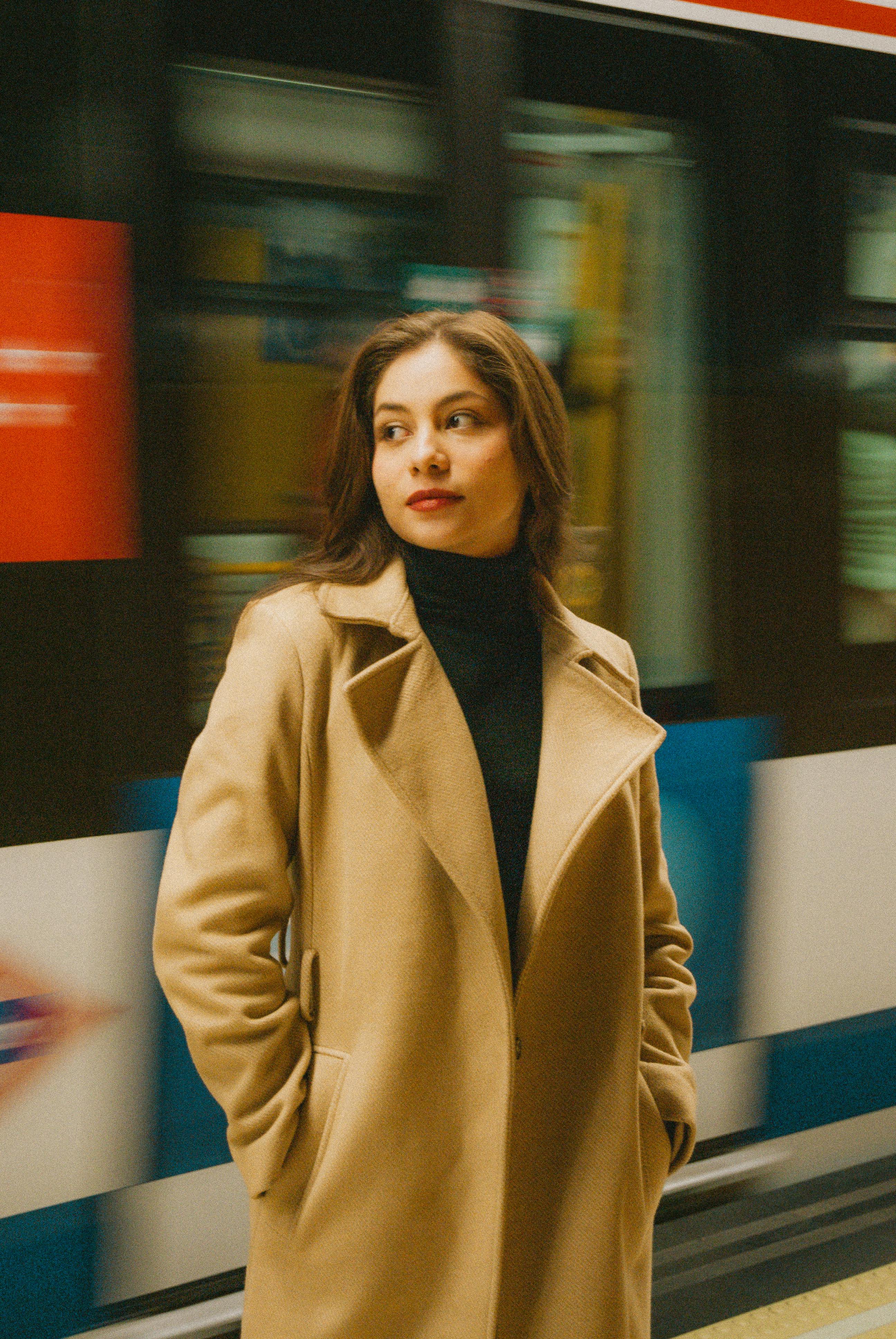 Elegant woman in a beige coat standing at Madrid subway station with blurred train passing by.