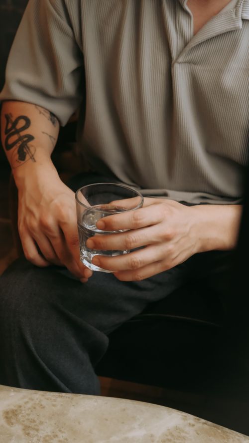 Free A seated man with tattooed arm holding a glass of water, indoors setting. Stock Photo