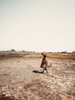 A lone African woman walks through an arid landscape with a wide skirt, carrying a basket on her head, under a bright summer sky.
