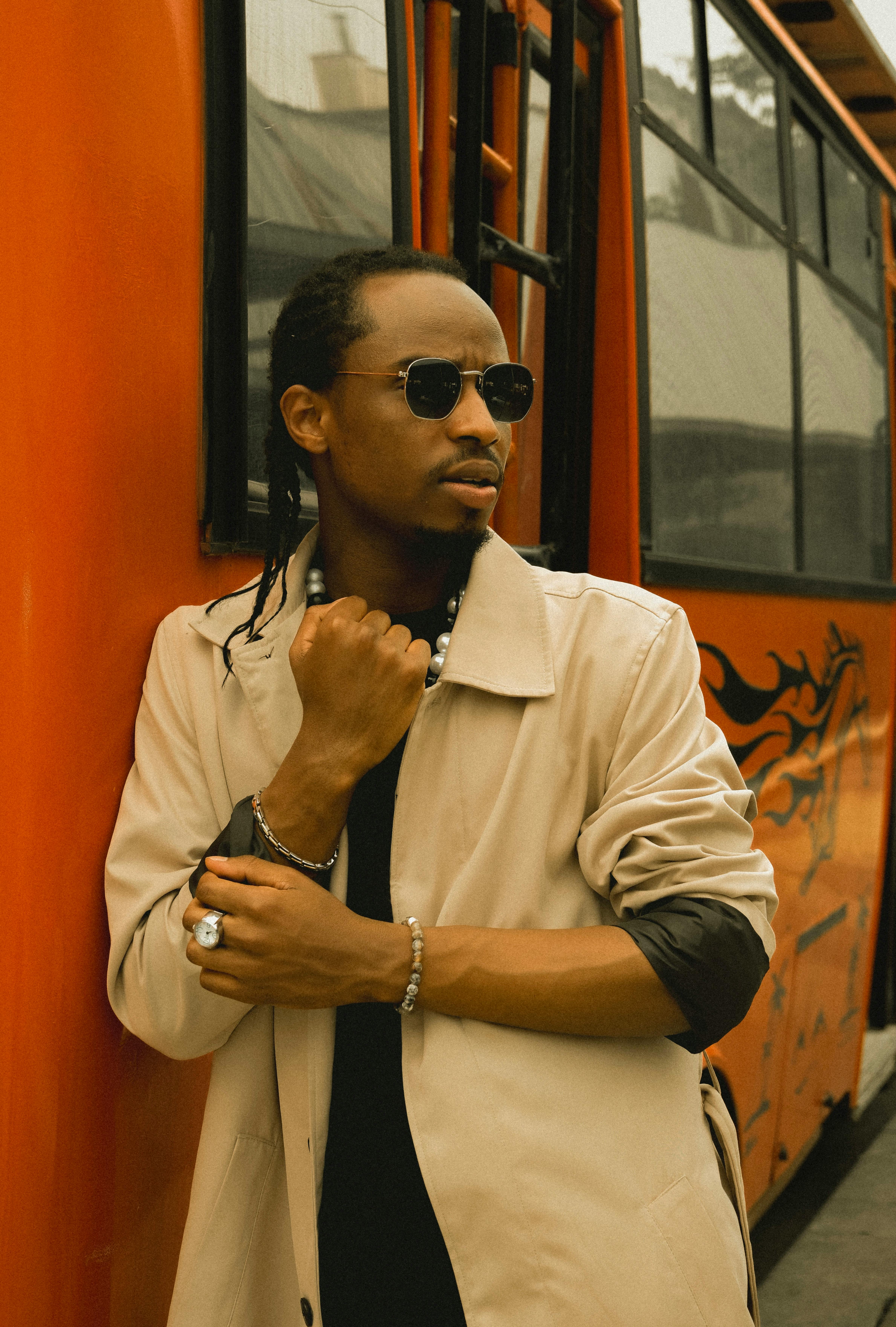 Fashionable man wearing sunglasses and coat standing by a vibrant orange bus in an urban setting.