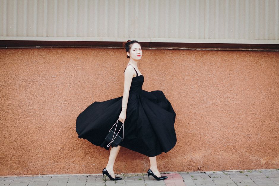 Stylish woman in black dress and heels walking confidently by a brick wall, showcasing elegance.