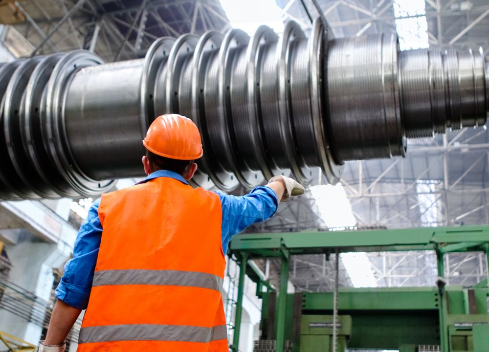 Worker in a high-visibility vest and hard hat guiding a large machined steel shaft in a factory bay.