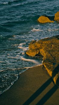 Golden sunlight casts shadows on a rocky beach with foamy waves at sunset.