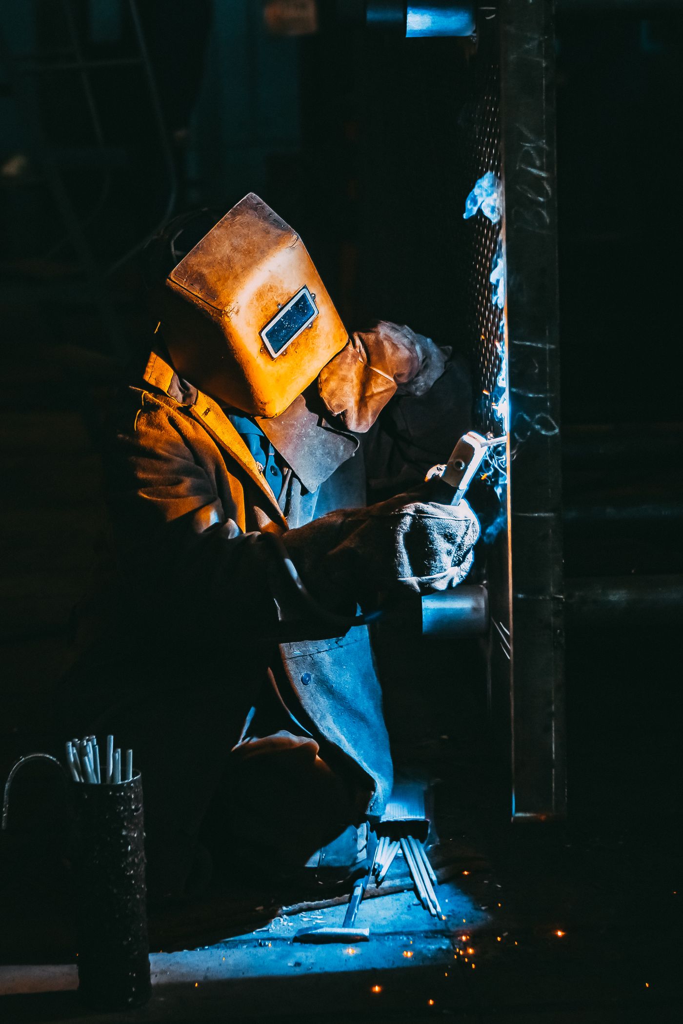 Welder in a full mask laying a weld on a steel upright, arc light throwing shadows across the bay.