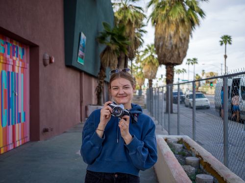 Free A woman in a blue hoodie holds a camera, smiling on a sunny urban street with palm trees. Stock Photo