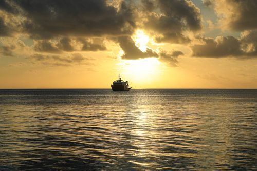 Free A serene sunset casts a warm glow over a tranquil ocean, featuring a silhouetted ship on the horizon. Stock Photo