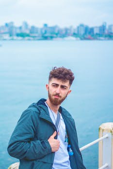 A man in a teal jacket poses by the waterfront with a city skyline in the background.