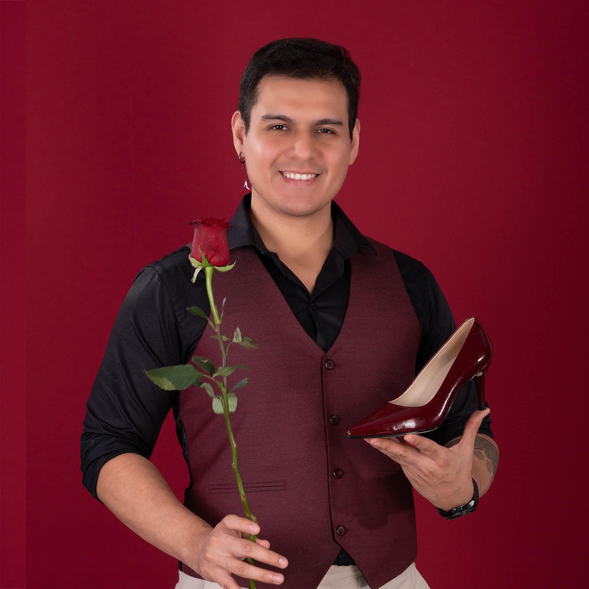 A smiling young man holding a red rose and a matching high heel shoe against a red studio background.
