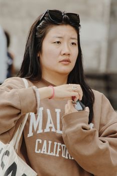 A young woman adjusts her sunglasses while wearing a casual sweatshirt outdoors.