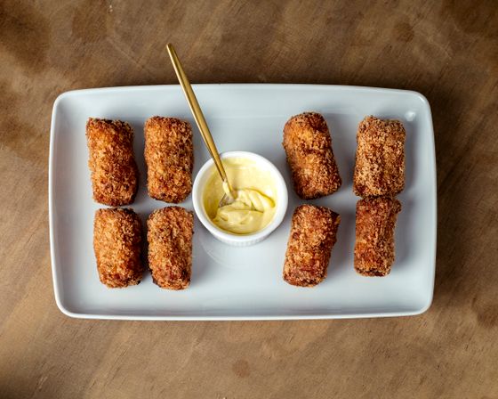 Eight dark golden crumbed croquetas on a white rectangular platter around a bowl of aioli