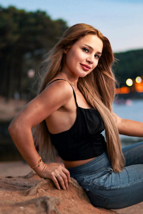 Free A young woman enjoys a serene summer evening by the water, showcasing a relaxed and stylish look. Stock Photo