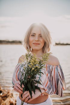 一位年轻女子面带微笑，手持鲜花，站在阳光明媚的湖畔