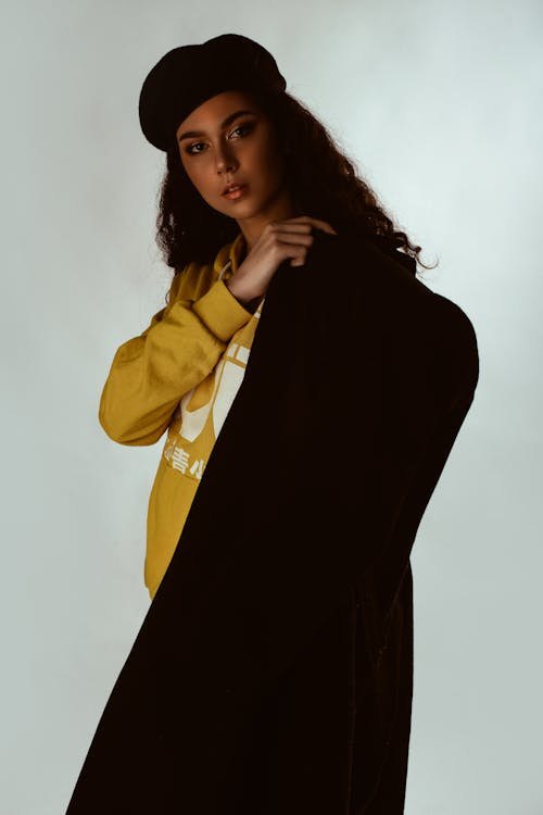 Photo of Woman in Yellow Hoodie, Black Beret Hat, and Black Coat Posing In Front of White Background