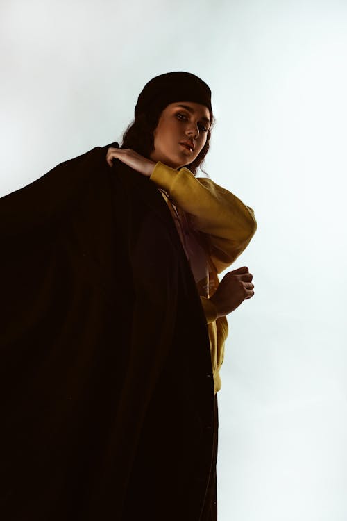 Photo of Woman in Yellow Hoodie, Black Beret Hat, and Black Coat Posing In Front of White Background