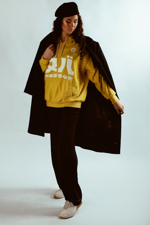 Free Photo of Woman in Yellow Hoodie, Black Beret Hat, and Black Coat Posing In Front of White Background Stock Photo