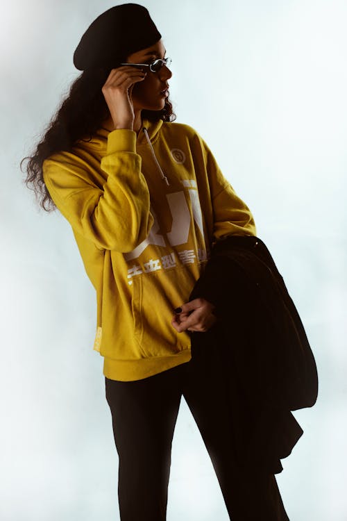 Photo of Woman in Yellow Hoodie, Black Beret Hat Posing In Front of White Background While Carrying Black Coat