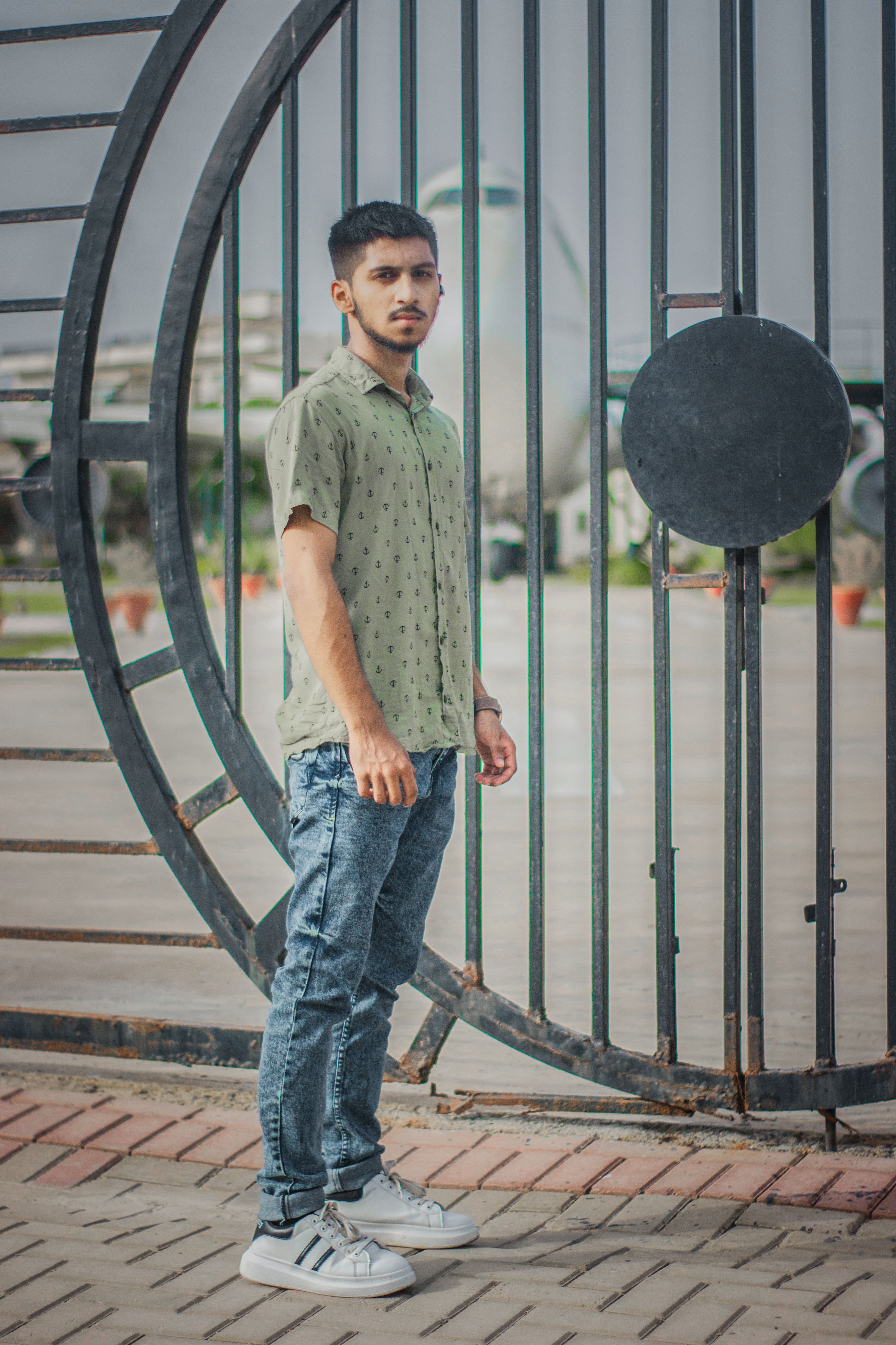 A young adult stands casually by a large urban fence, showcasing a modern fashion style.
