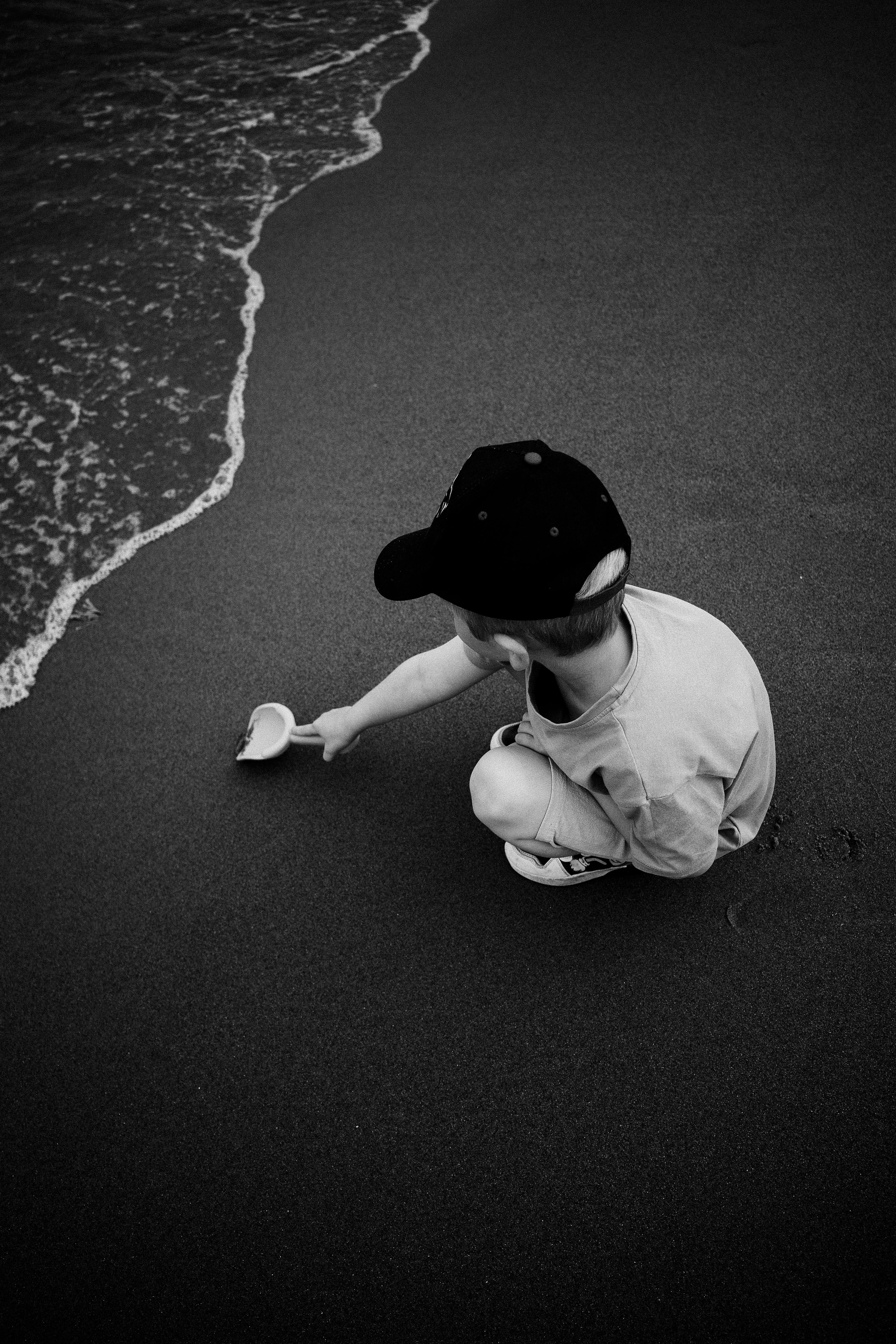 Child plays with a shovel on the sandy beach, near ocean waves, in a serene black and white photo.