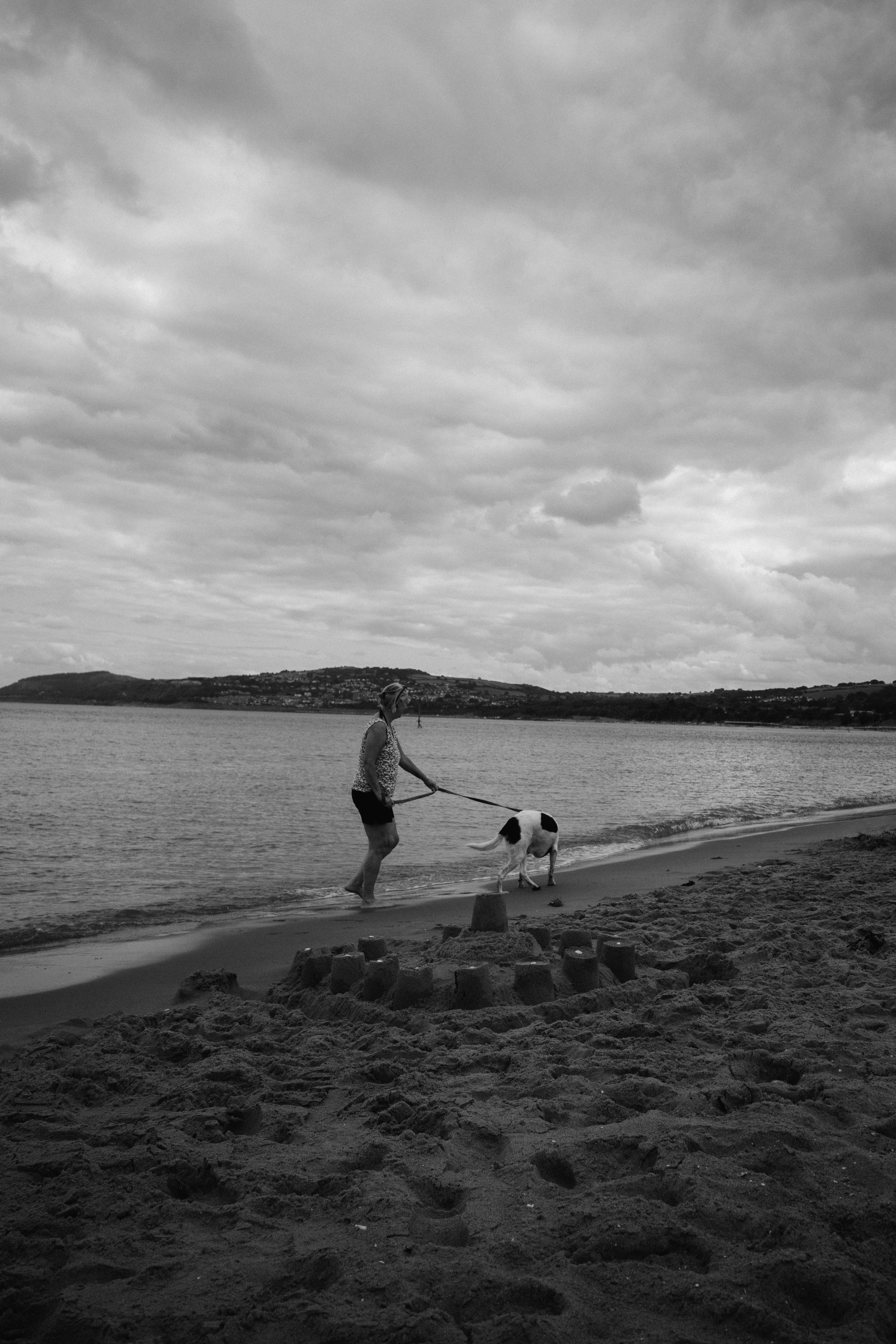 A person walks a dog on a quiet beach as clouds gather overhead, capturing solitude and reflection.