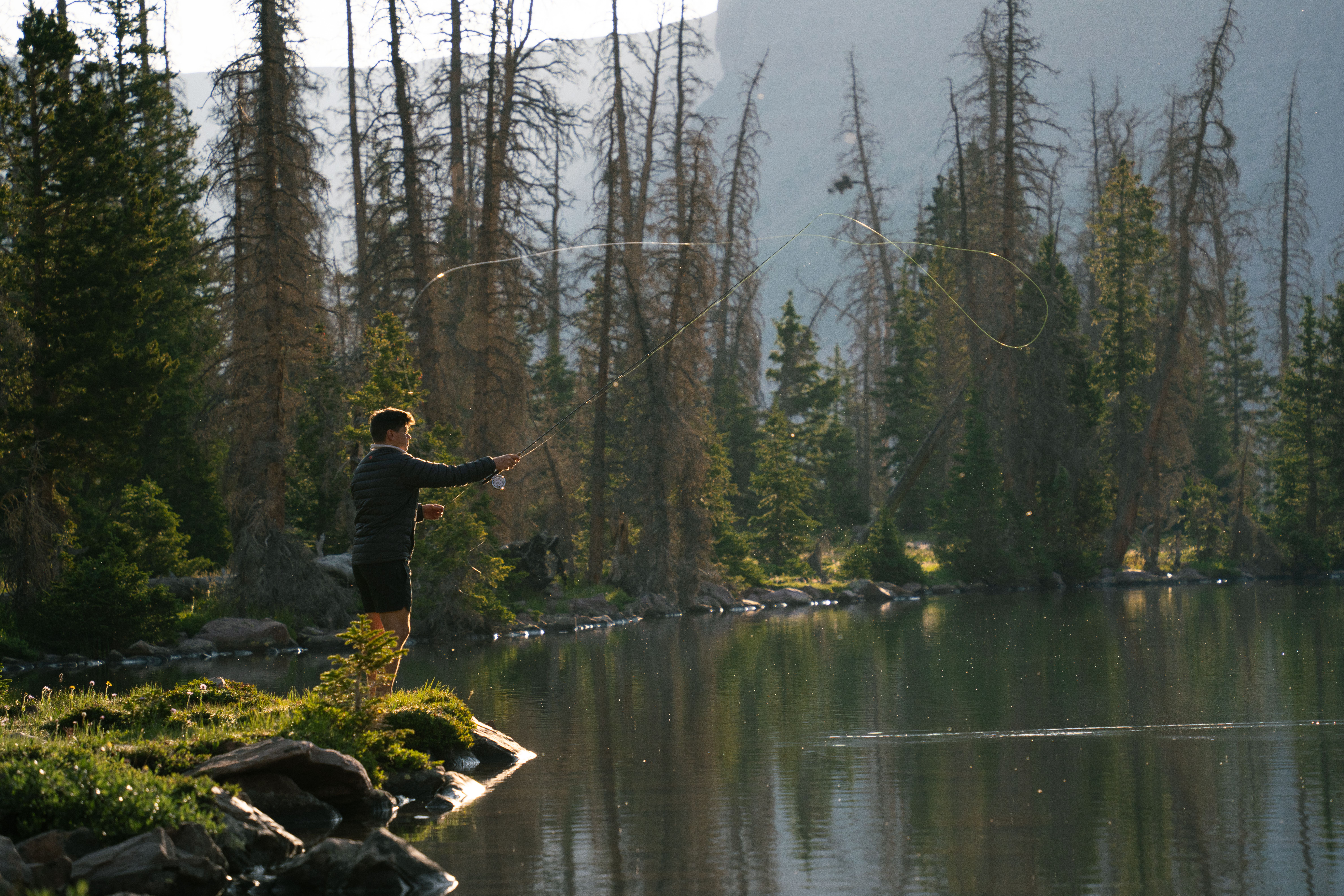A man fly fishing at a tranquil alpine lake in Utah's wilderness during dawn.