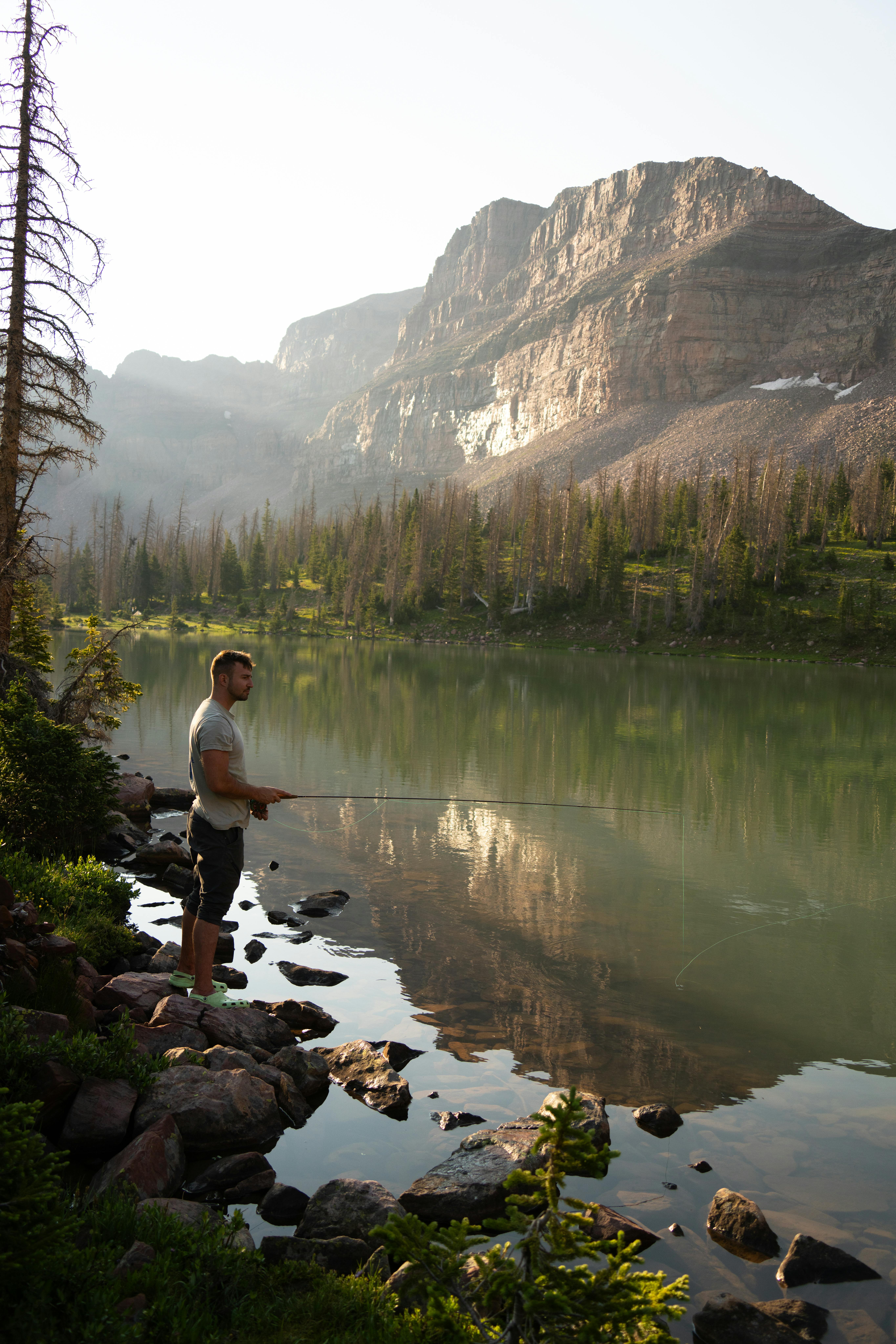 A man fly fishing by a calm mountain lake with reflection.