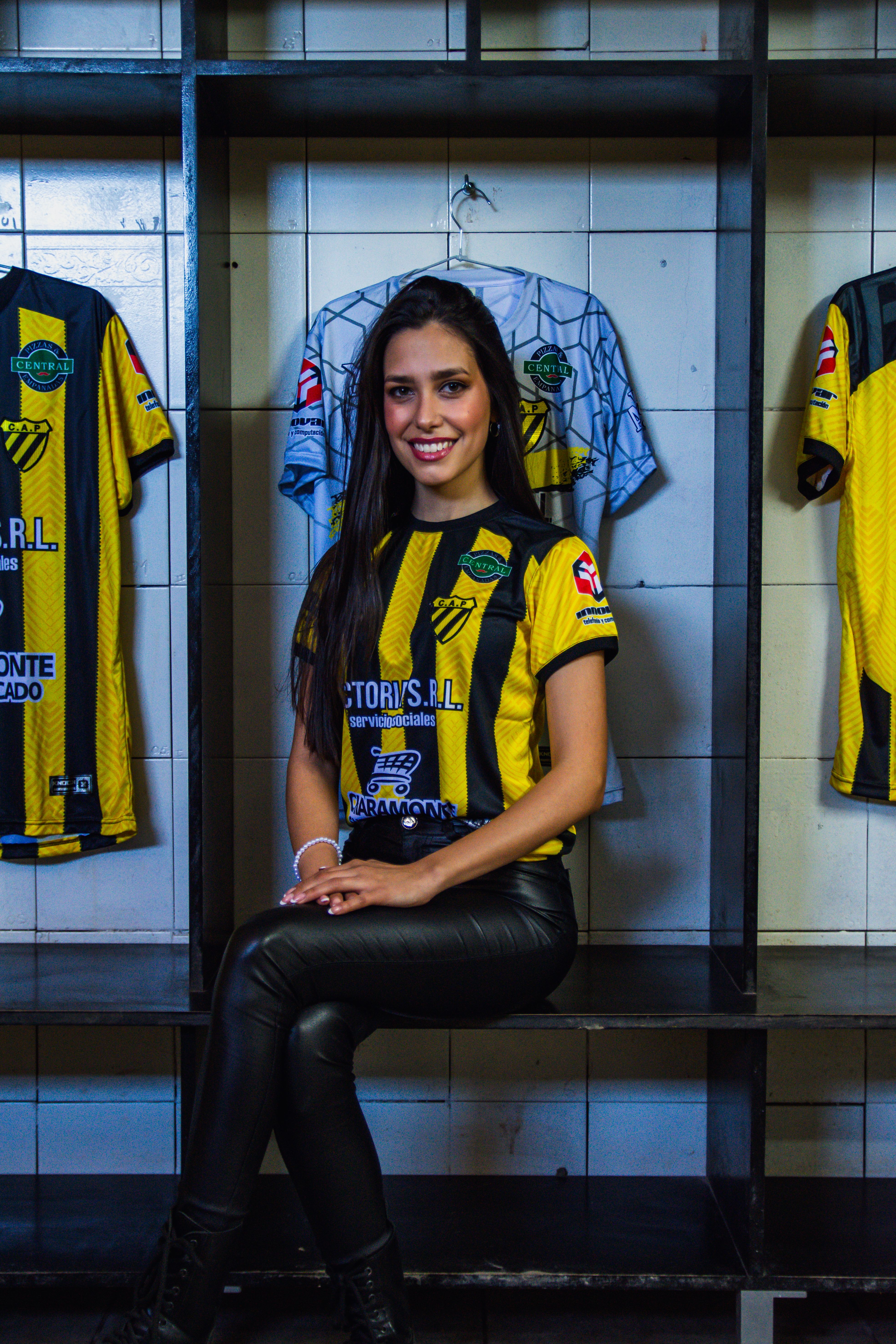A woman posing in a soccer jersey inside a locker room, radiating confidence.