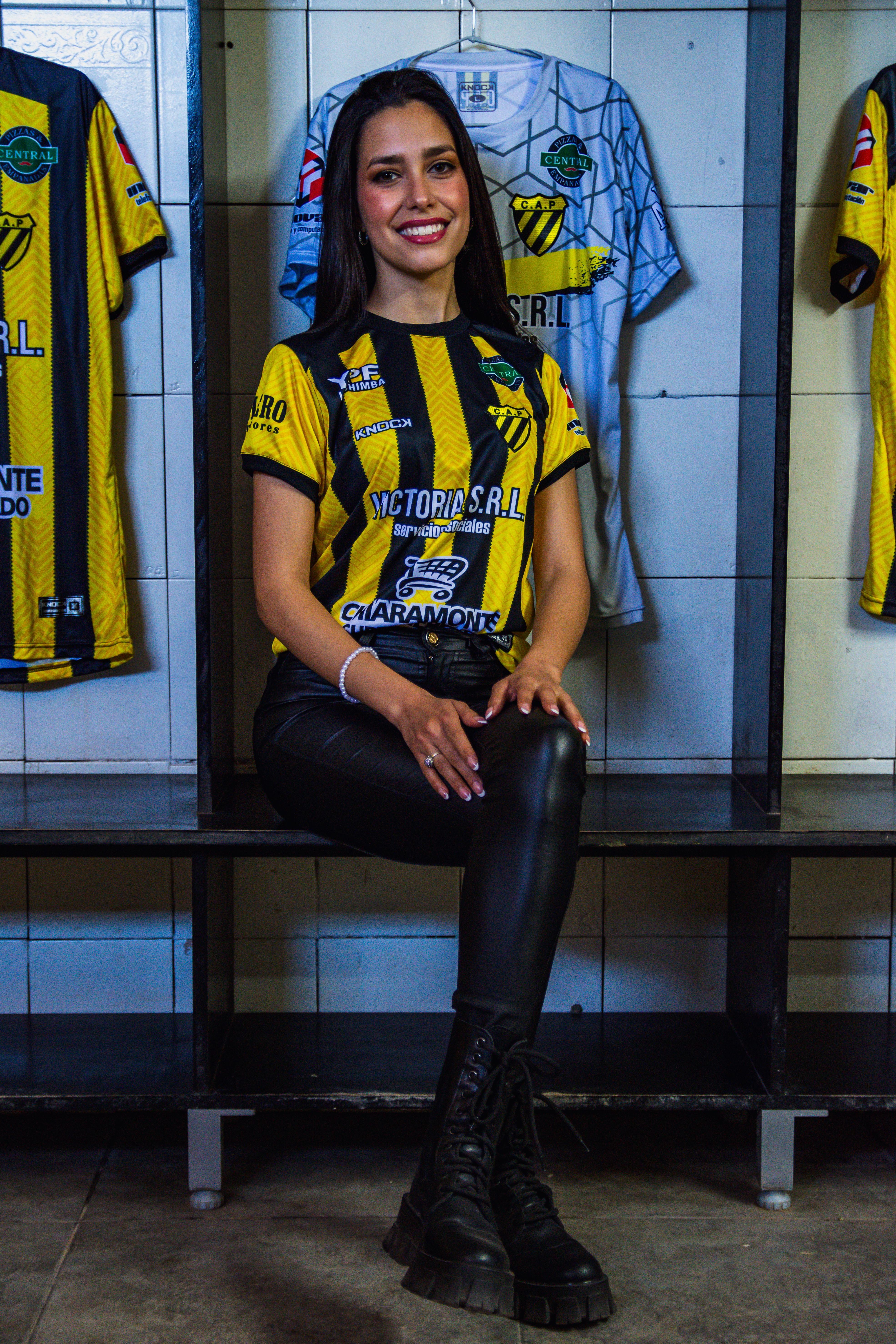 A woman in a yellow and black soccer jersey sits inside a locker room, exuding confidence.