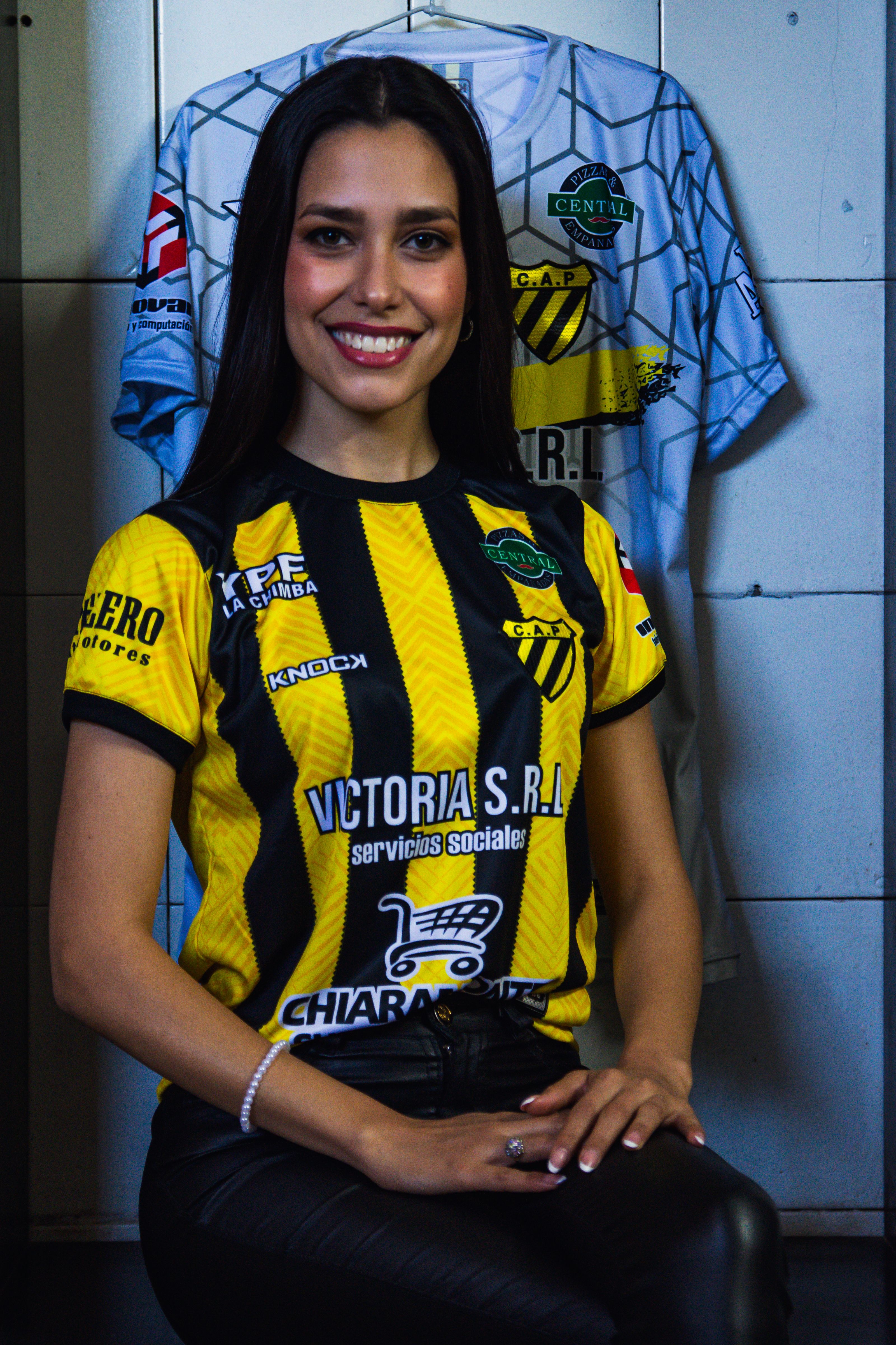 A smiling young woman wearing a soccer jersey sitting indoors, exuding confidence and style.