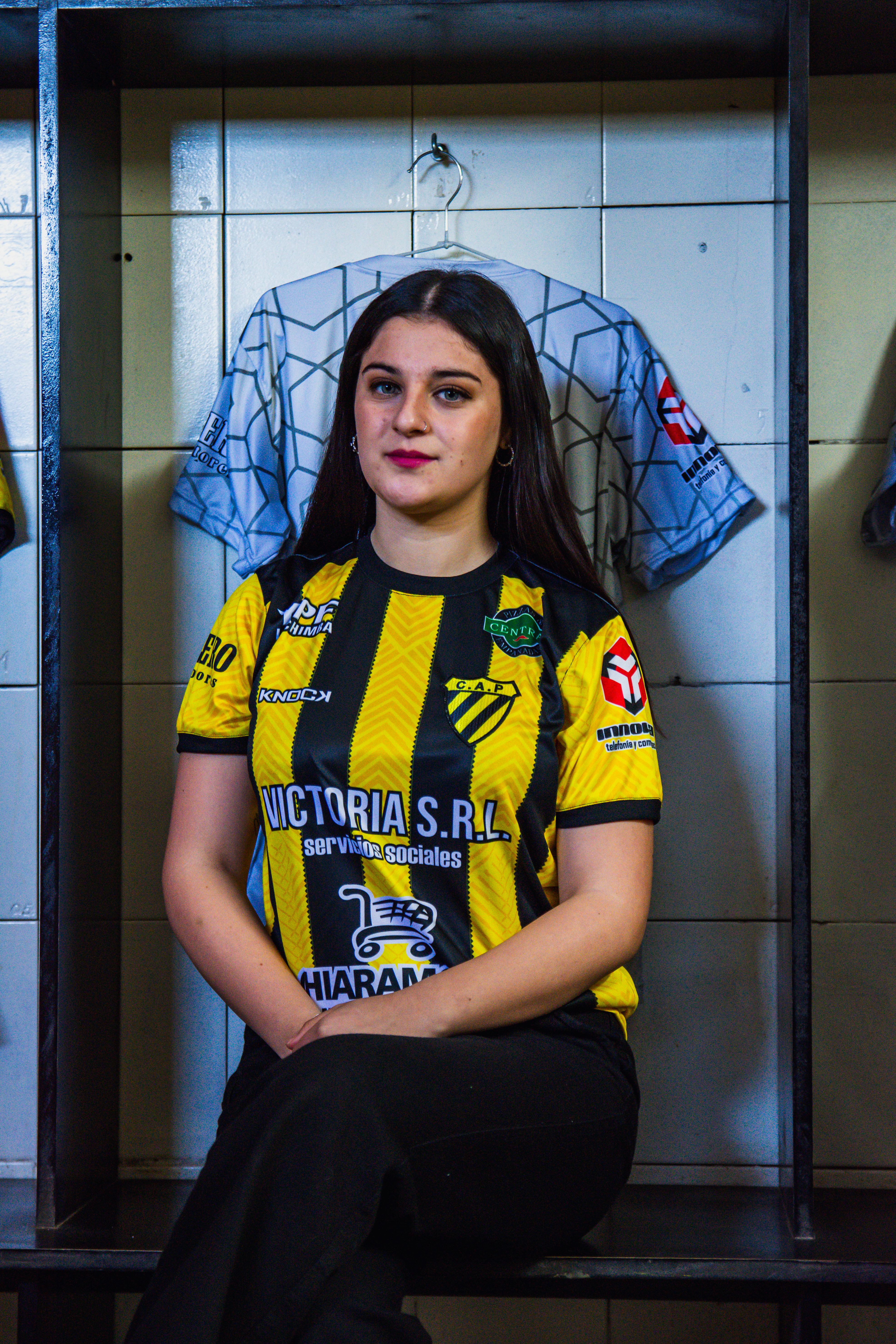 Portrait of a young woman in a soccer jersey sitting indoors, conveying team spirit.