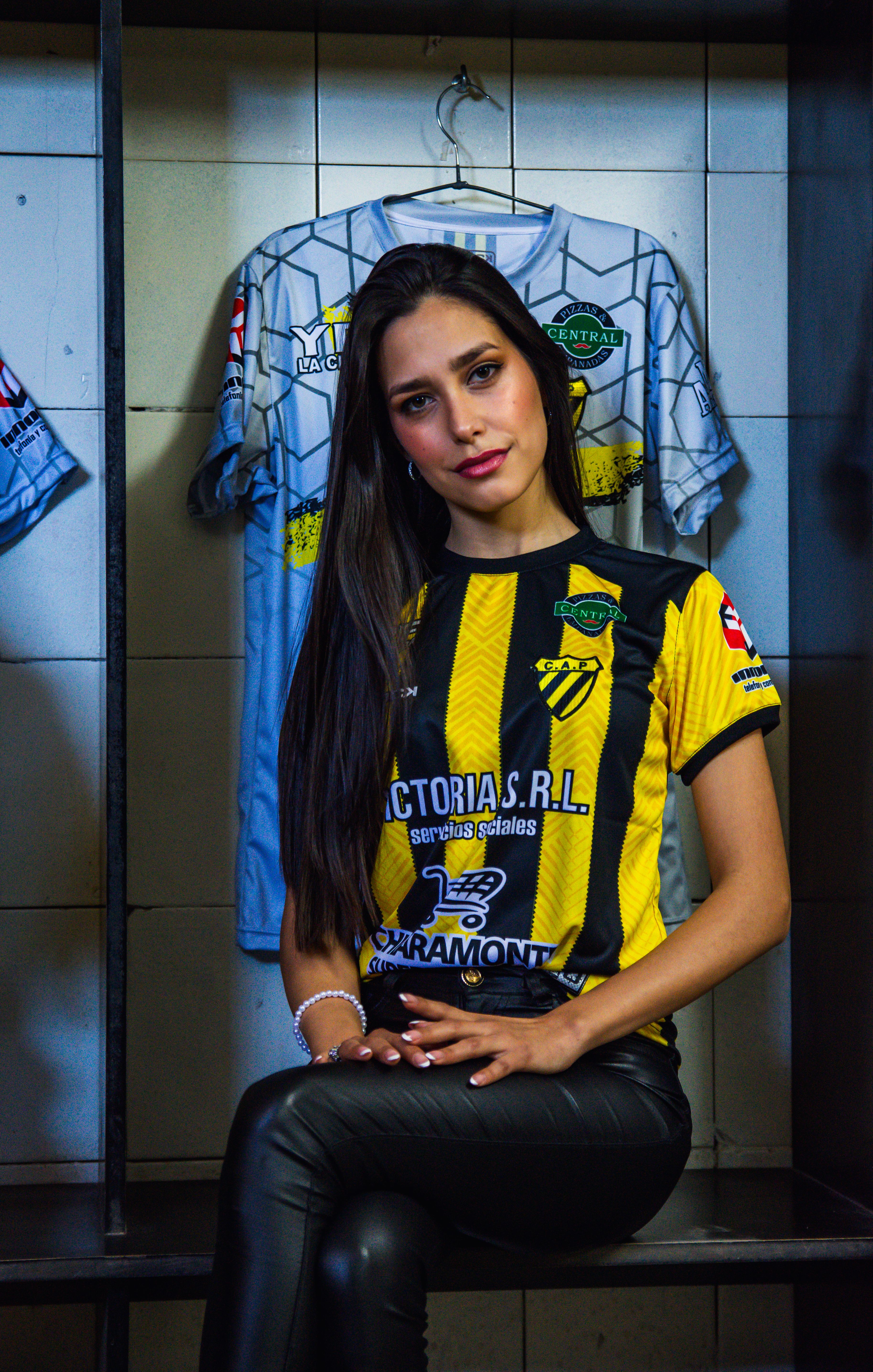 Confident young woman poses in a soccer jersey in a locker room setting.