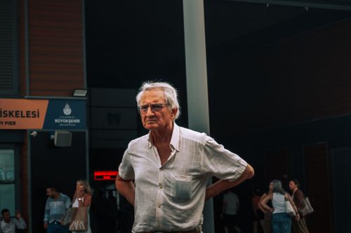 Free Elderly man stands with hands on hips in a busy urban setting with people in the background. Stock Photo