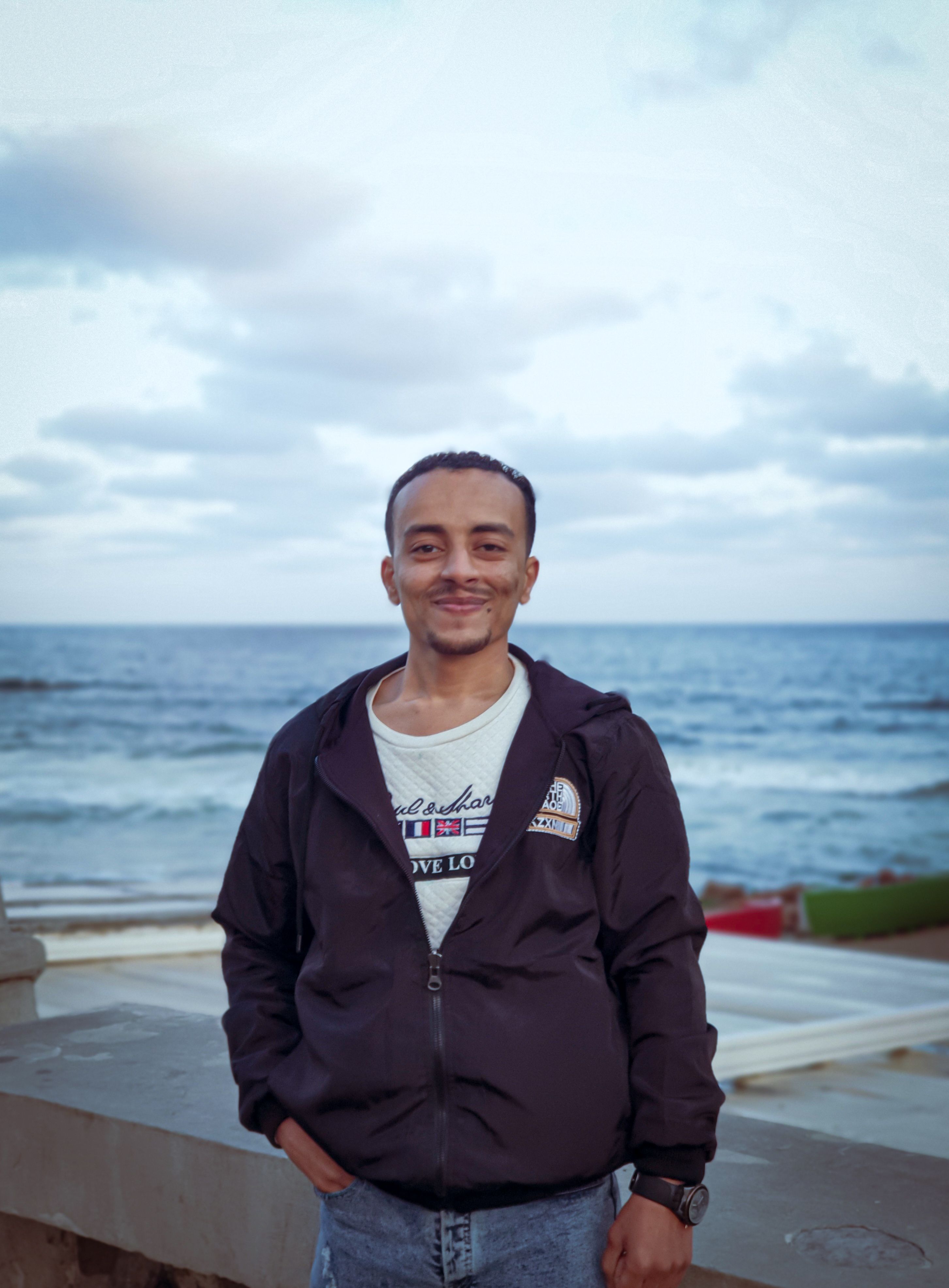 A smiling man enjoys a peaceful moment by the sea in Alexandria, Egypt, under a cloudy sky.