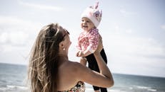 一位快乐的母亲抱着她微笑的婴儿，背景是风景优美的海滩