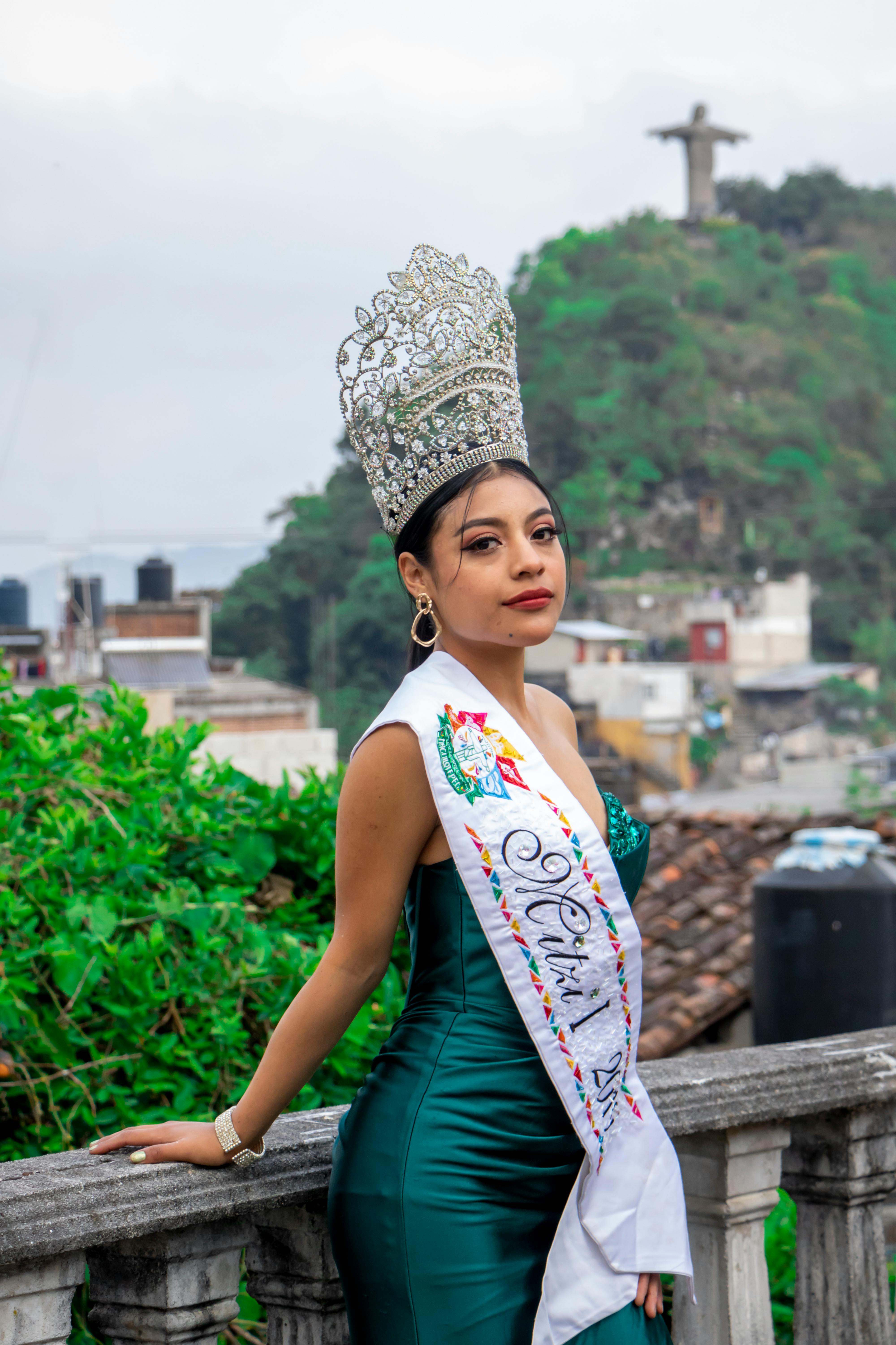A young beauty queen in an elegant gown and crown, posing outdoors with a cityscape backdrop.