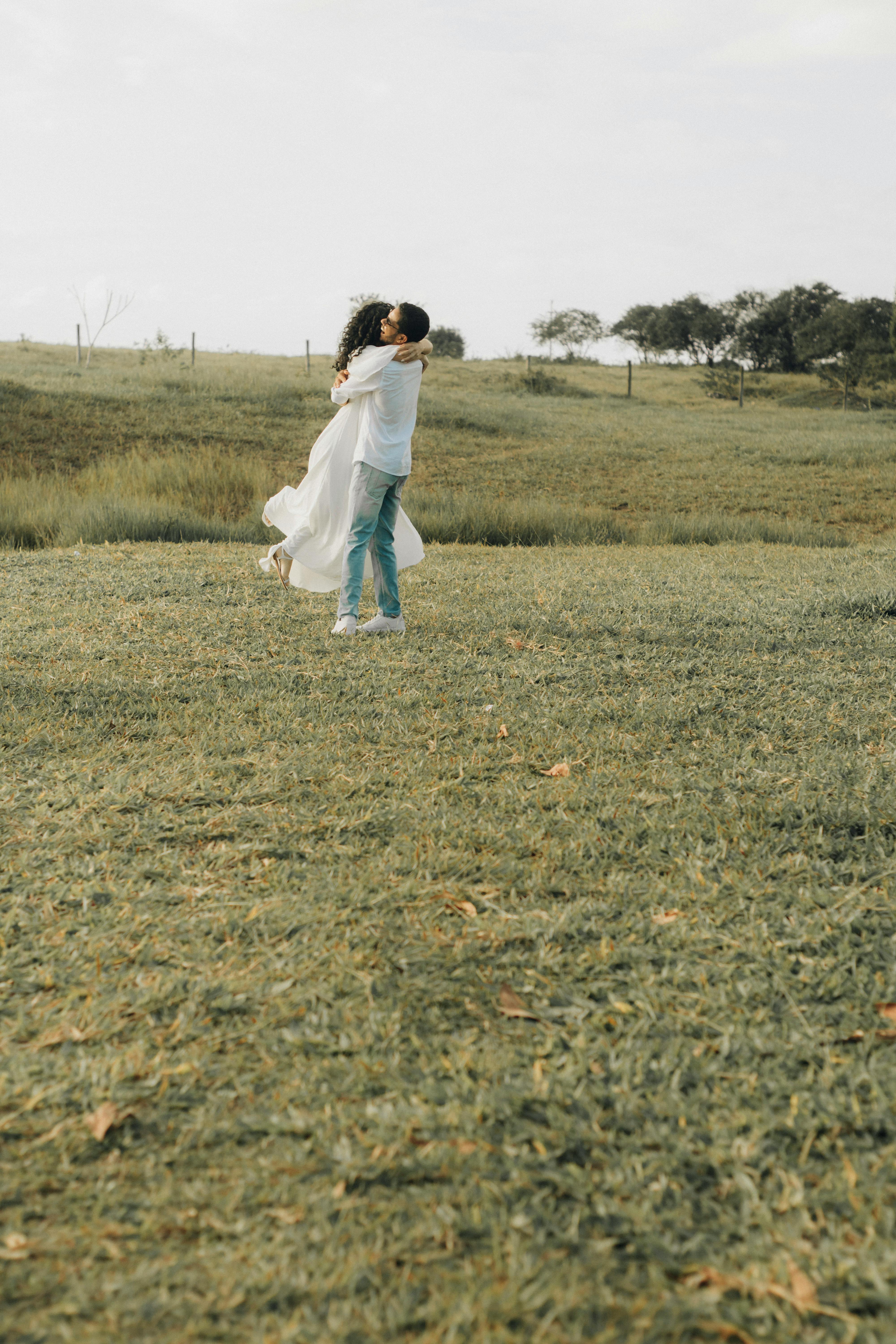 A couple in casual attire embraces in a scenic outdoor field, capturing a moment of joy.
