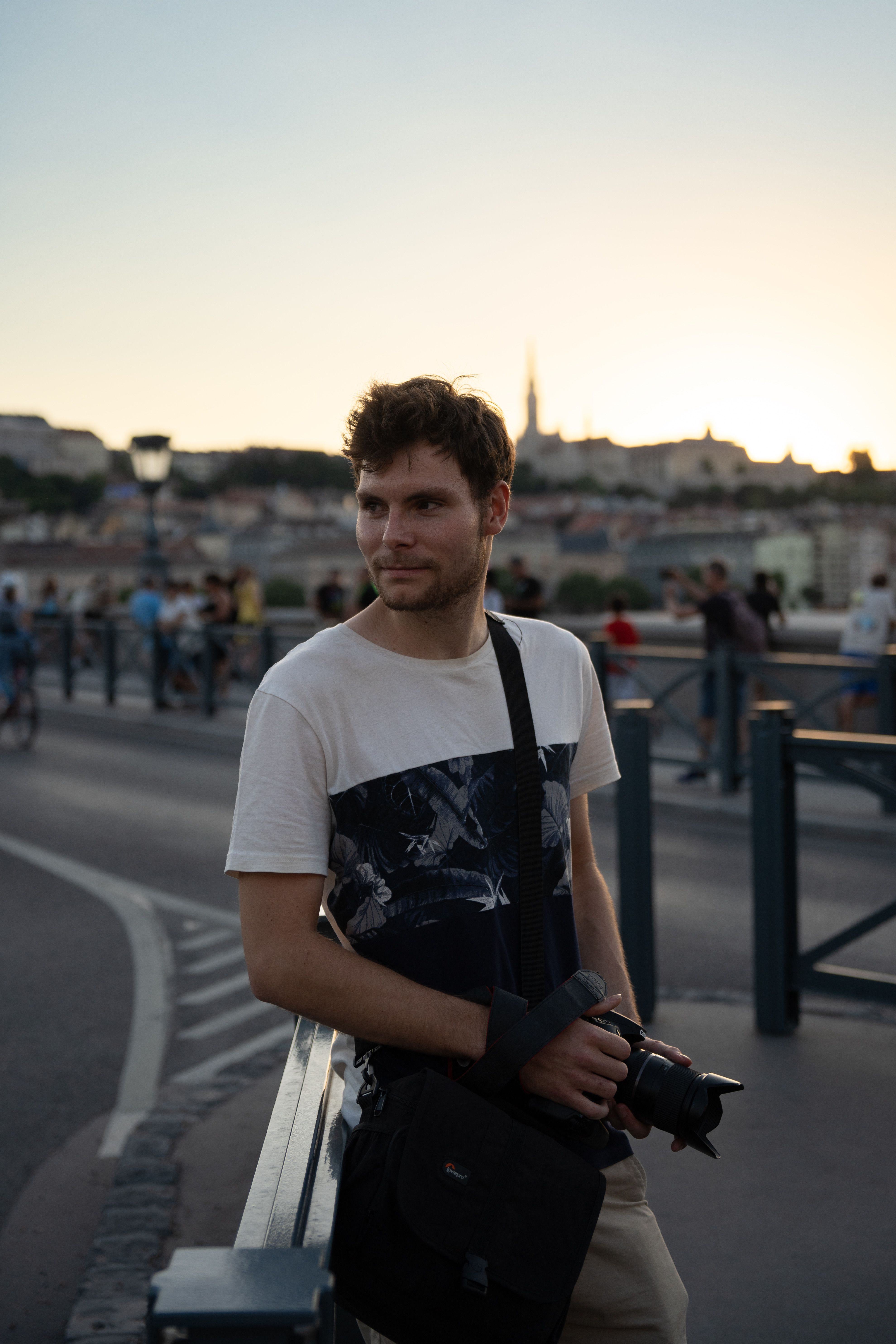 A thoughtful young man with a camera rests on a bridge during sunset in an urban setting.