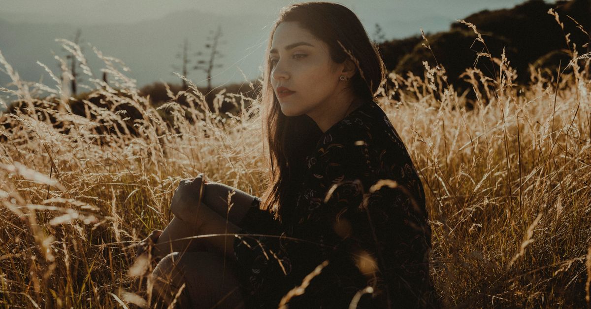 person sitting alone in a quiet rural field, hands on knees, looking thoughtful yet burdened