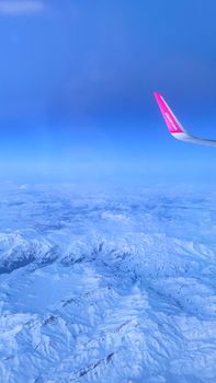 Aerial shot of snowy mountains from an airplane window with a bright pink wingtip against a clear blue sky.