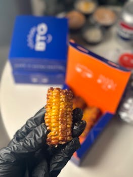 Close-up of grilled corn held in gloved hand with a takeout box in the background.