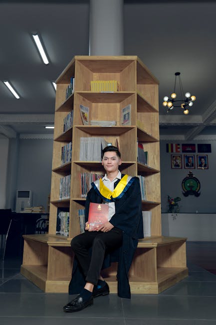 Young adult in graduation attire poses in university library, holding diploma.