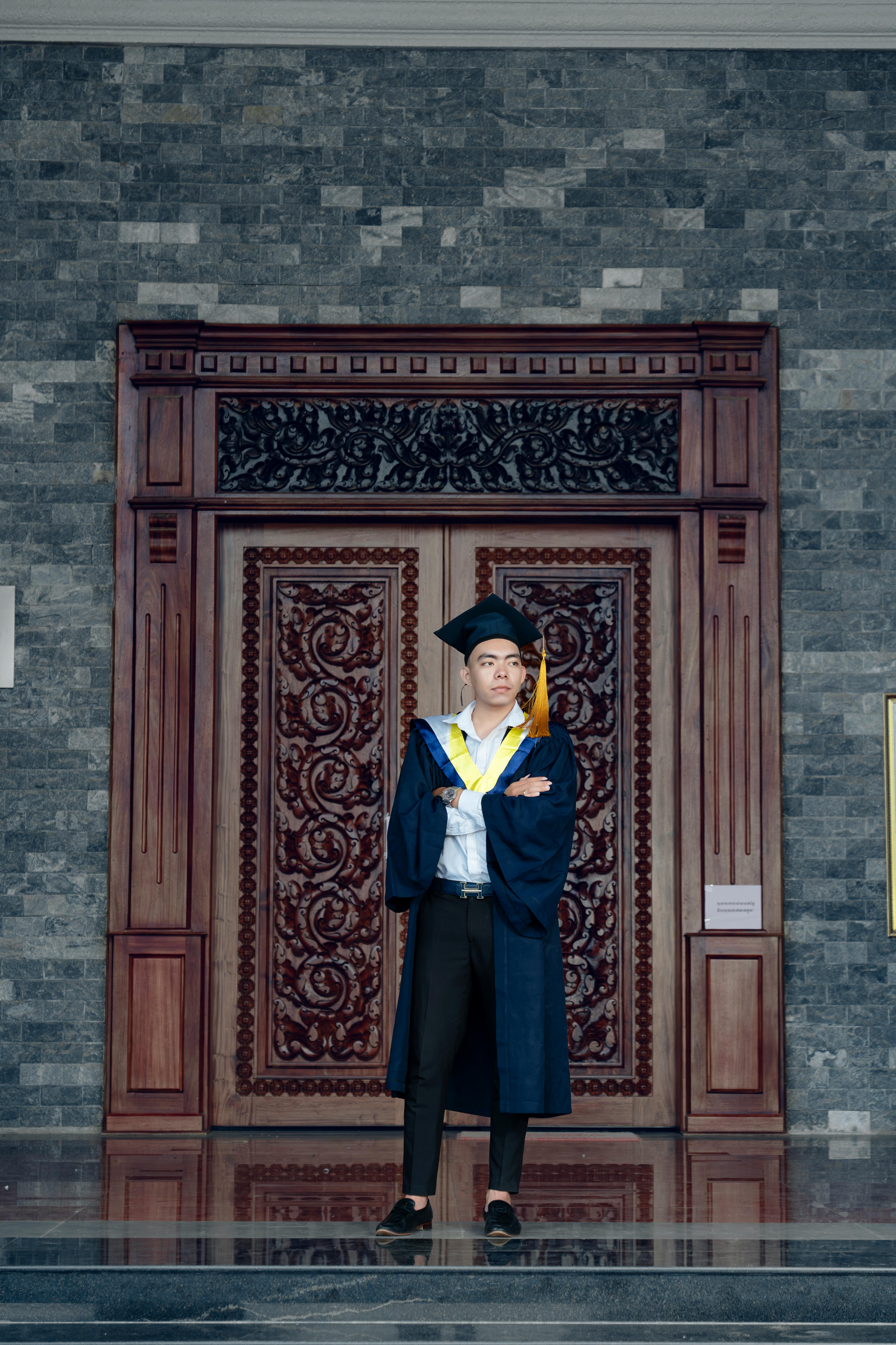 Young adult in graduation attire stands confidently in front of intricately carved wooden doors.
