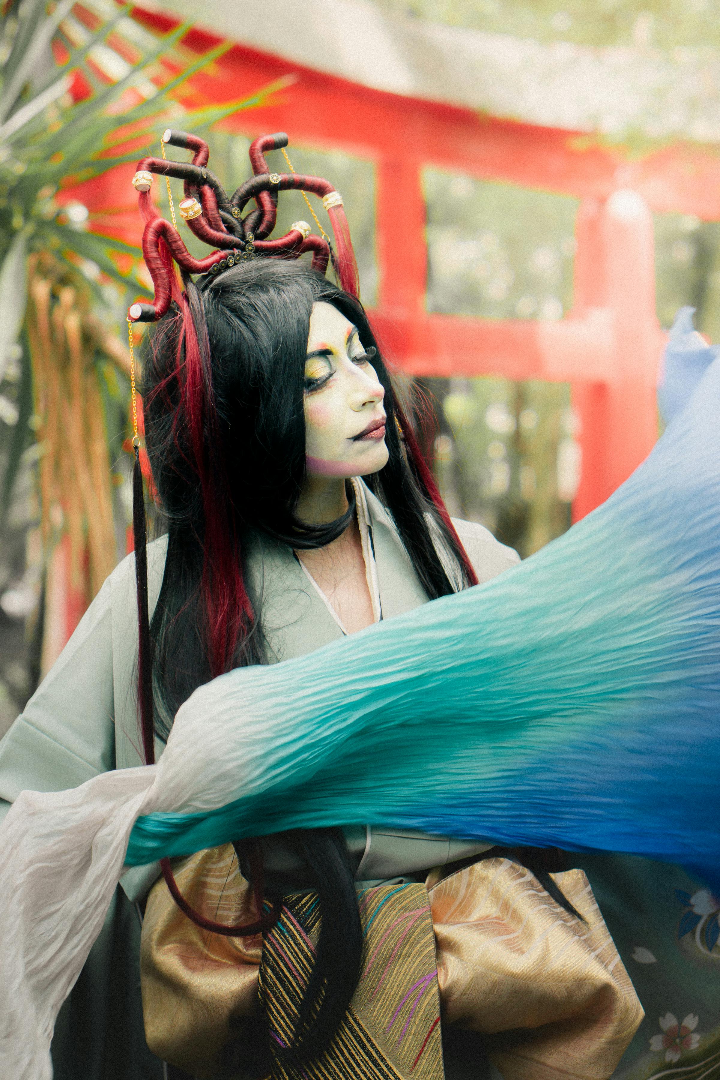 A woman in traditional Japanese kimono and elaborate hairstyle poses amidst vibrant colors outdoors.