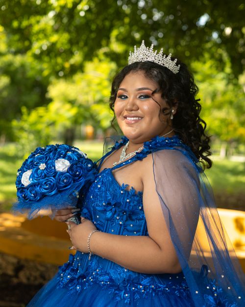 Free Smiling quinceañera in elegant blue dress with tiara and bouquet in sunny park. Stock Photo