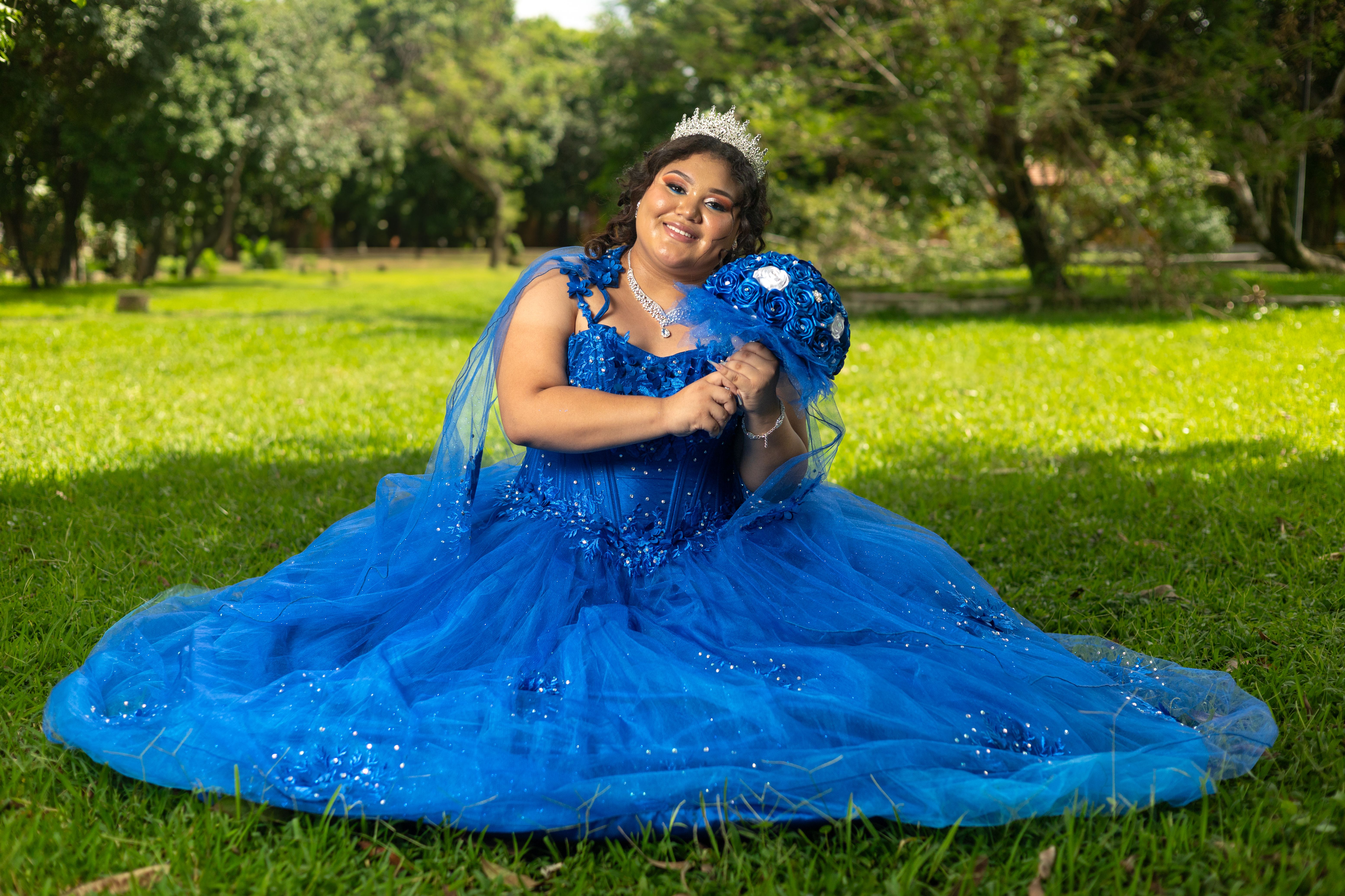 A quinceañera wearing a vibrant blue dress poses happily in a lush park setting.